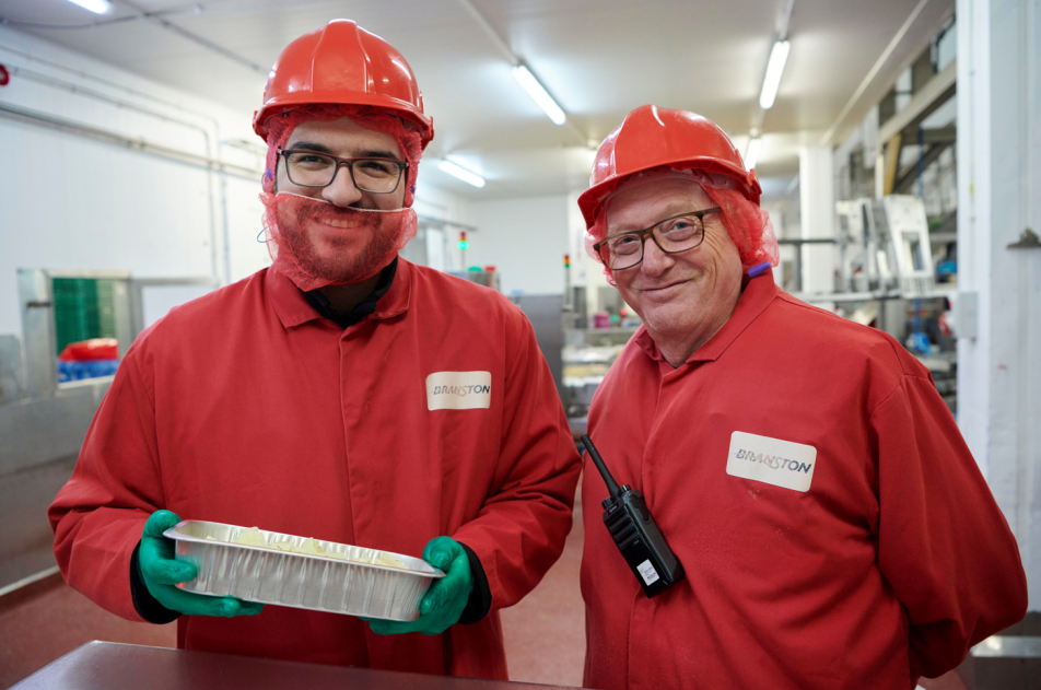 Two men in red overalls, hard hats and face covers holding mashed potato in foil tray